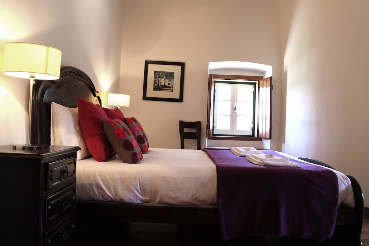 A hotel room with a bed, a nightstand, a lamp, a chair, a window, and a framed picture on the wall.