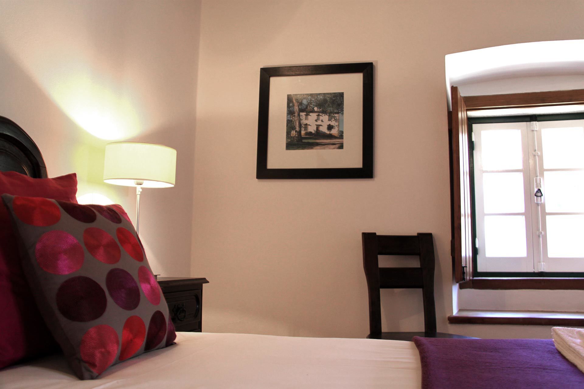 Een hotelkamer met een bed, een nachtkastje met een lamp, een lijst met een foto aan de muur, een stoel en een raam met uitzicht.