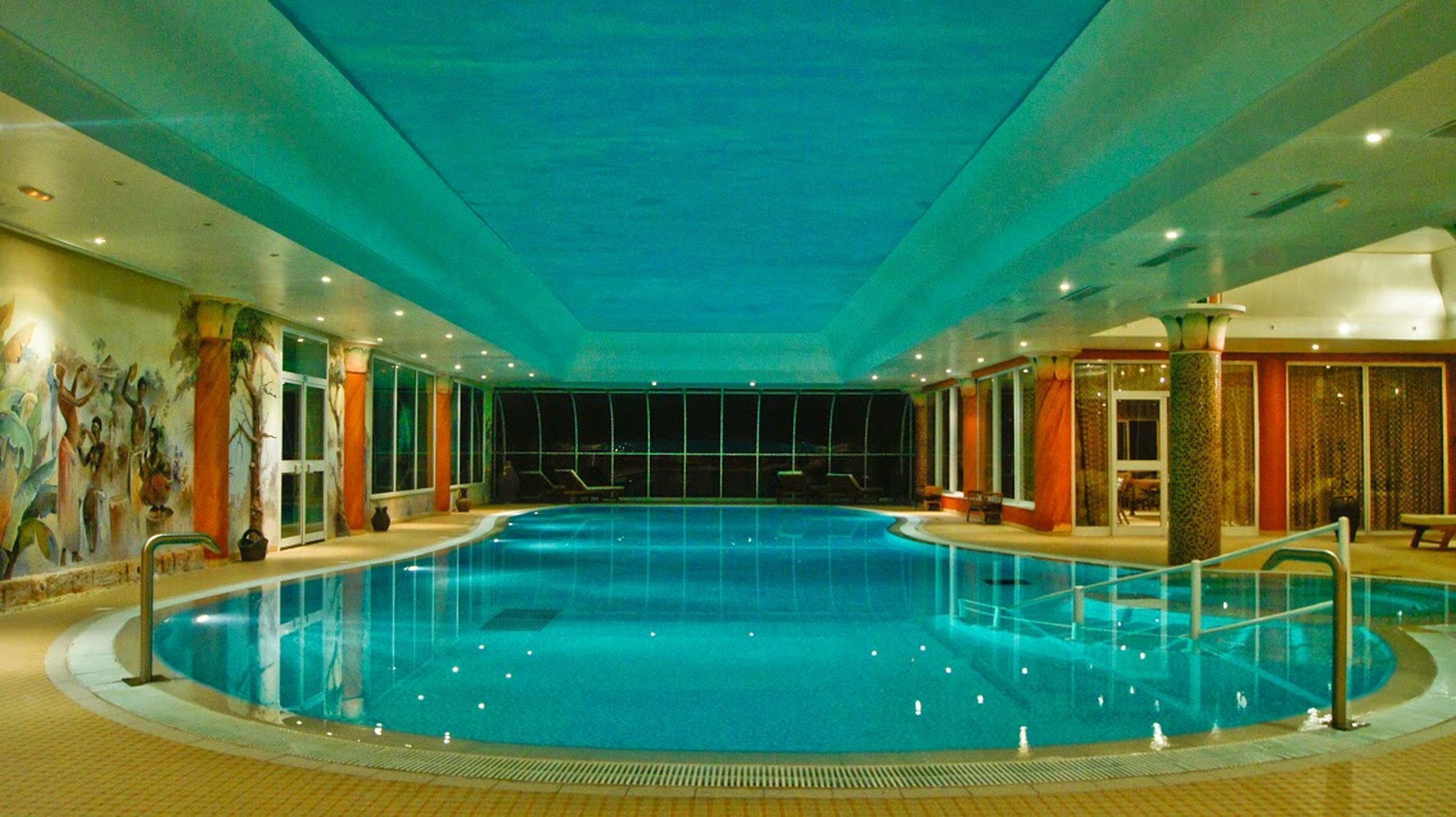 Una piscina interior con suelo de baldosas, zona de trampolín central y paredes con murales.