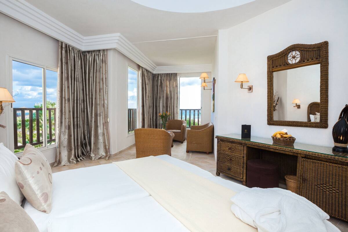 A spacious bedroom with a large bed, two armchairs, a small table, and a mirror.