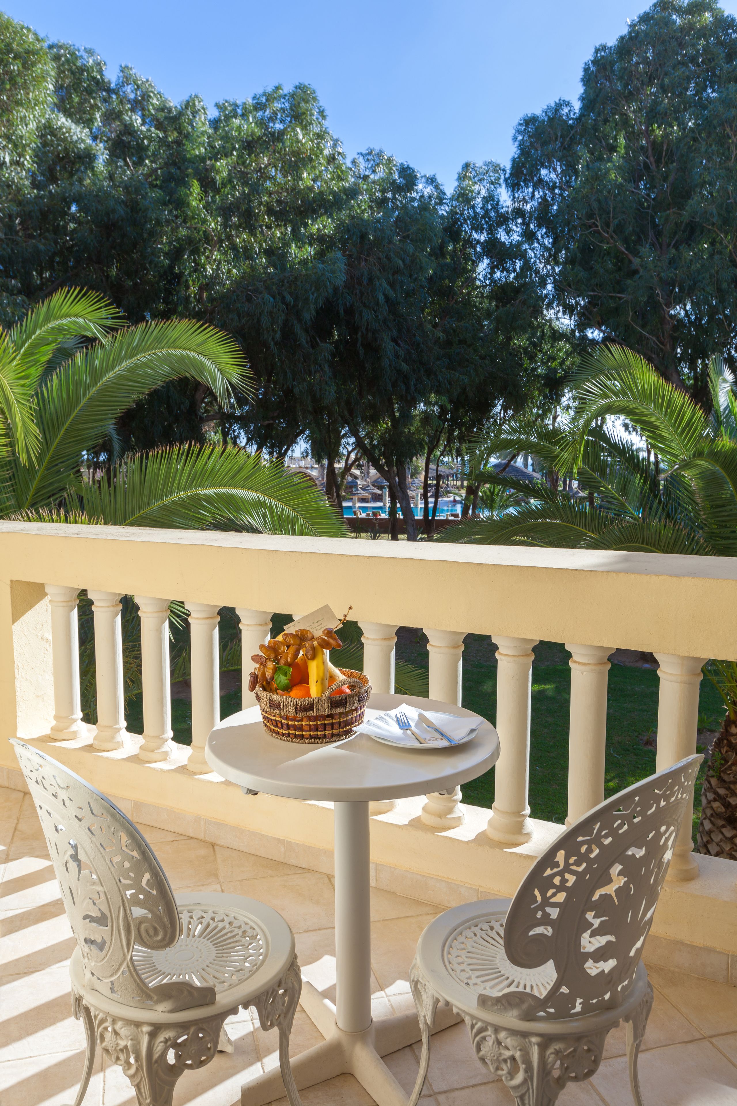 A balcony with a round table and two ornate chairs, a wicker basket with food items, and a view of trees and a pool in the background.