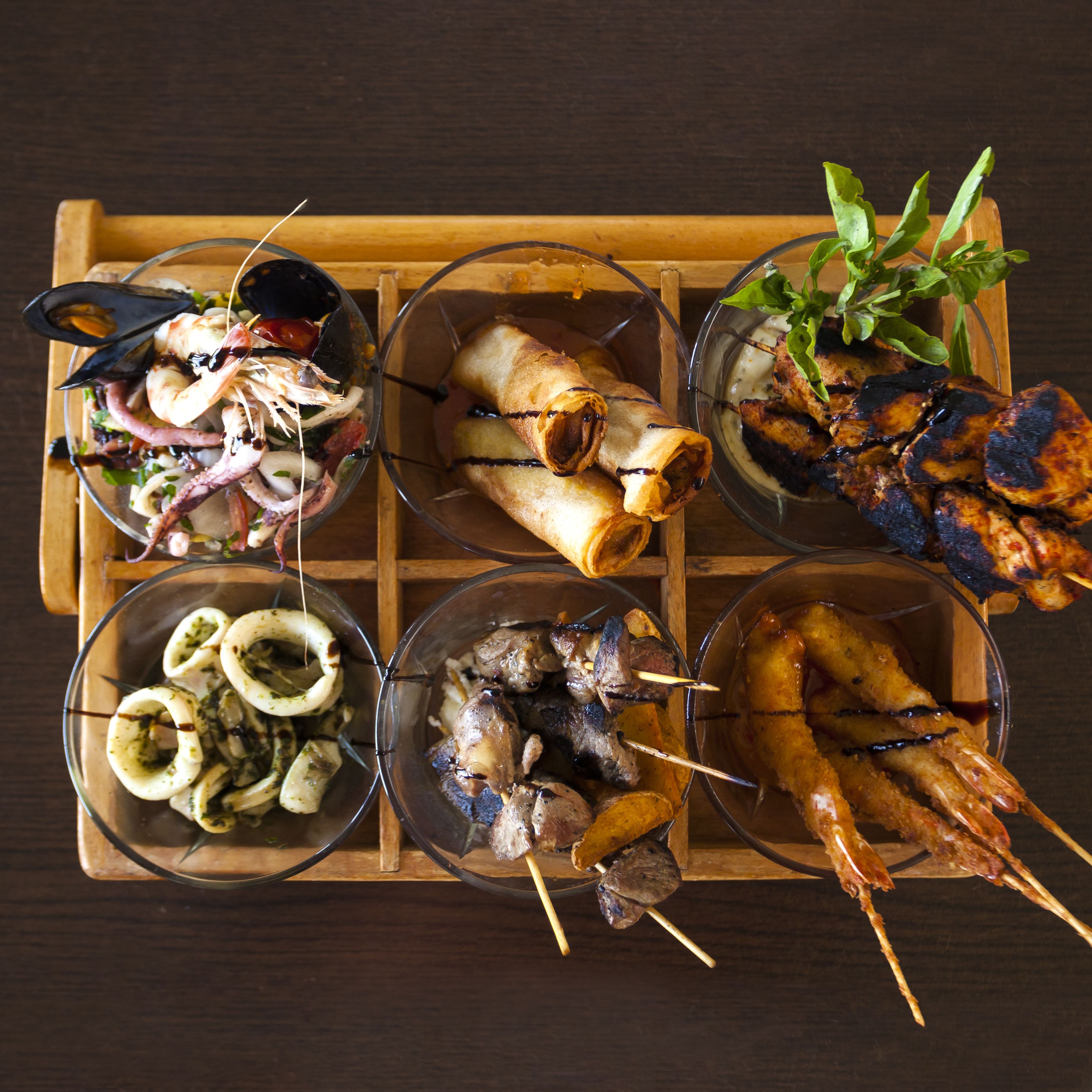 Una bandeja de madera con seis recipientes de vidrio que contienen diversos platos de mariscos.