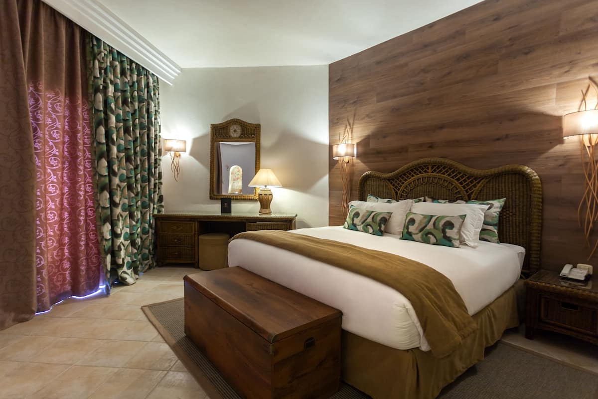 A bedroom with a wooden headboard, a bedside table with a lamp, a mirror, and patterned curtains.