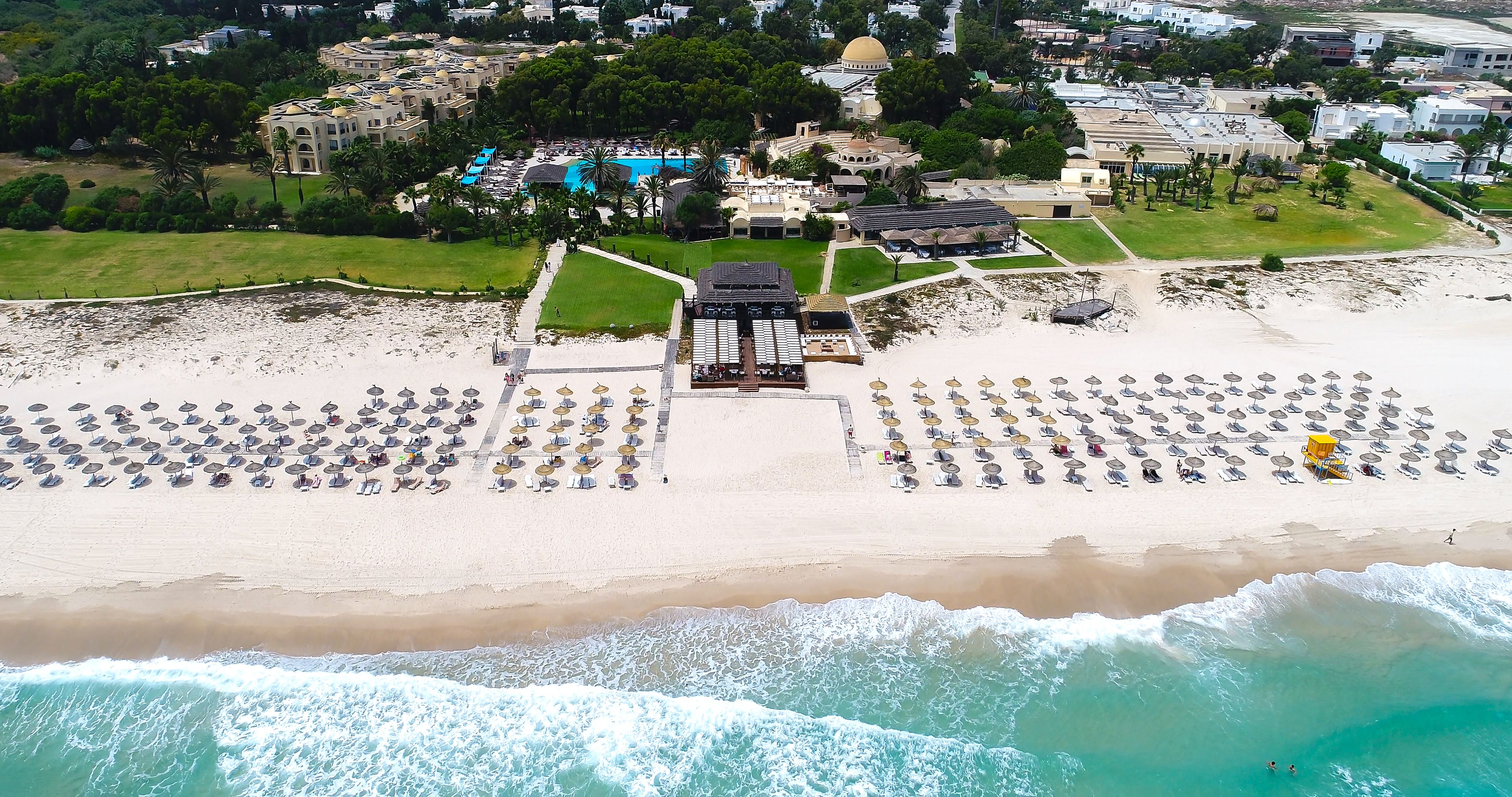 Vue aérienne d’un complexe hôtelier en bord de mer, avec une grande plage de sable, de nombreux parasols et un espace piscine.