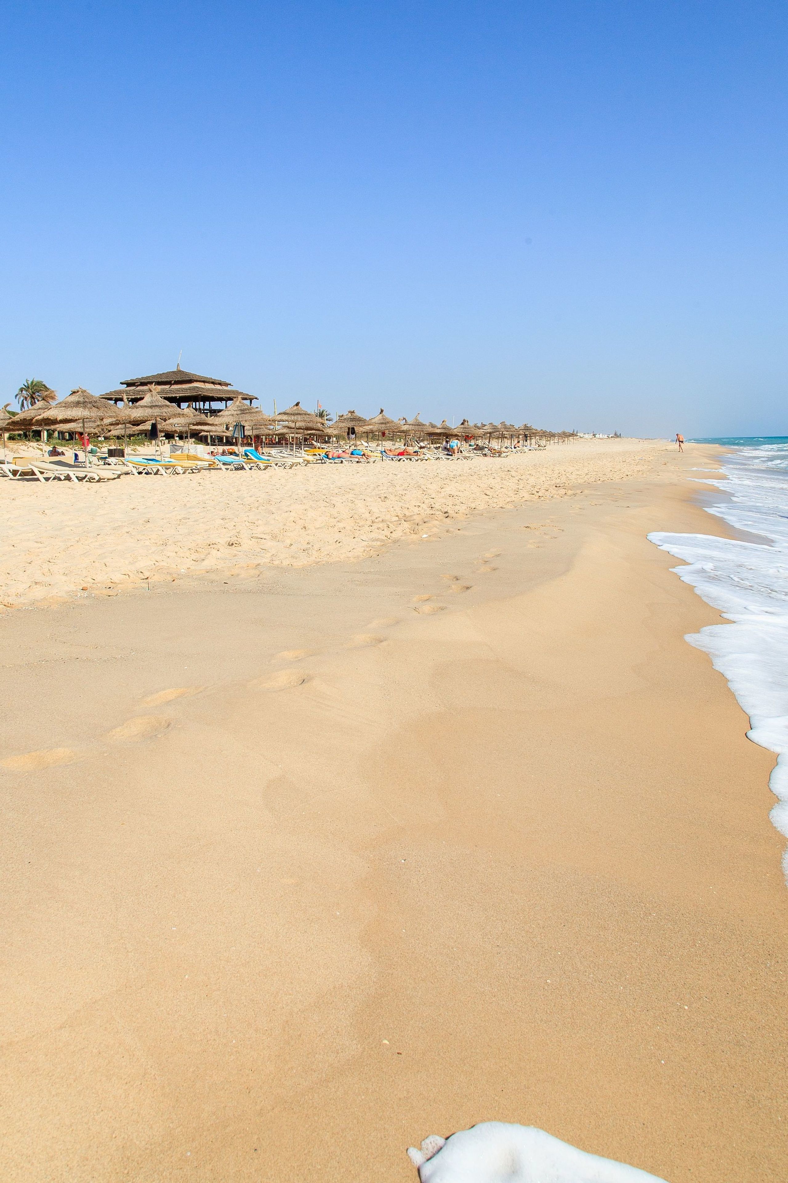 A beach with a row of thatched-roof huts, a sandy shore, and a clear blue sky.