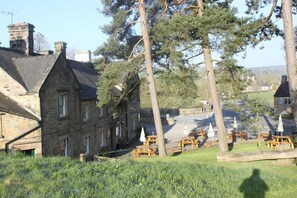 A stone building with a pitched roof, surrounded by trees and a grassy area with wooden picnic tables.