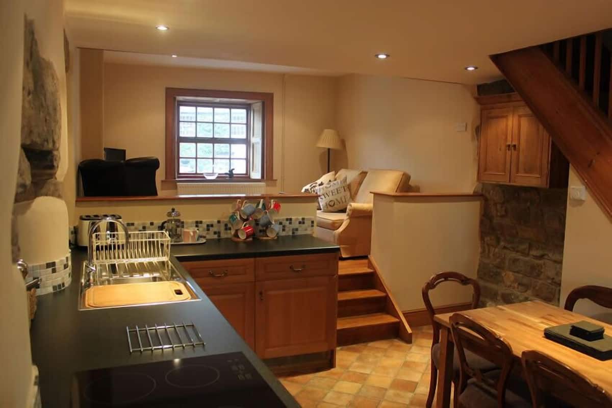 A kitchen with a black countertop, wooden cabinets, a window, a dining area with a table and chairs, and a staircase.
