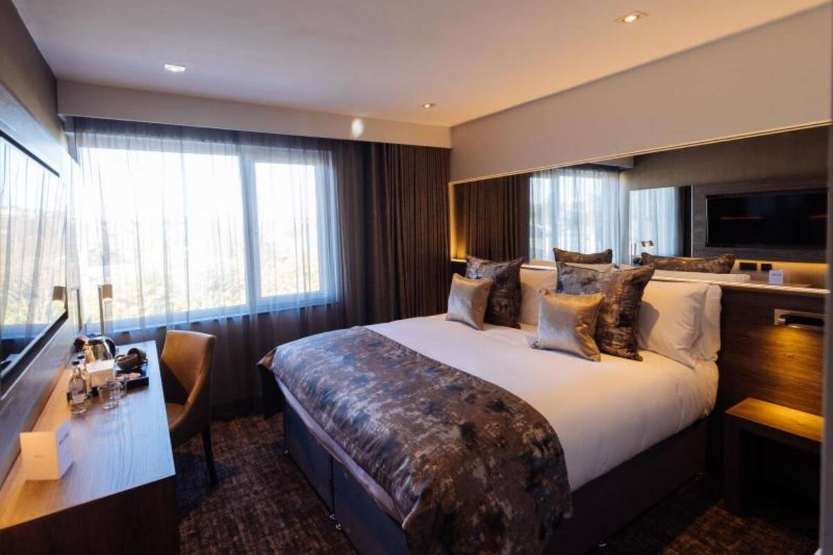 A modern hotel room with a large bed, a desk with a chair, a TV, and a large window with curtains.