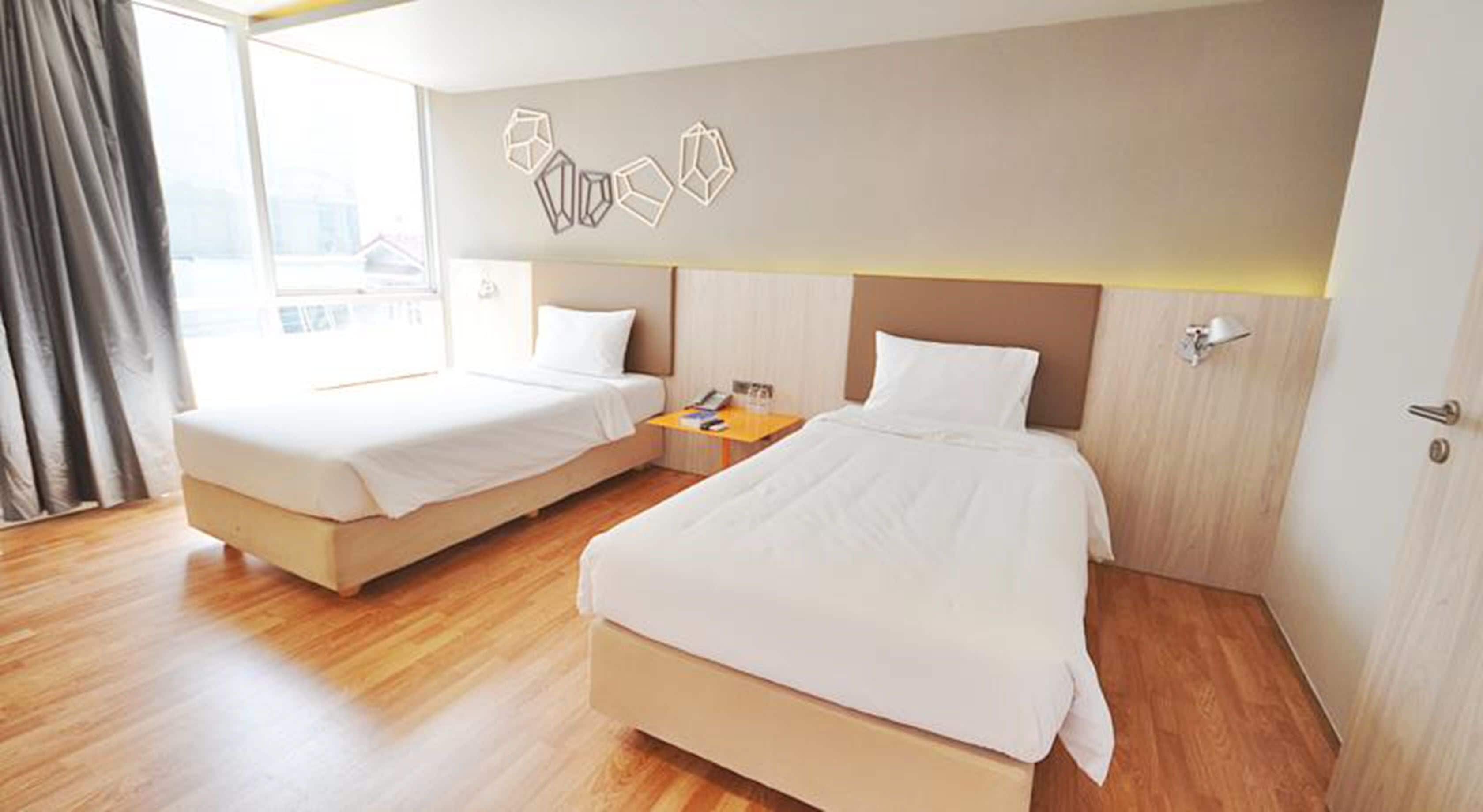 A hotel room with two beds, a wooden floor, a window with curtains, and a wall with geometric wall art.