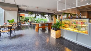 A café with wooden tables and chairs, a counter with a display case, and hanging plants. - The Heritage Hotels Bangkok (Bangkok)
