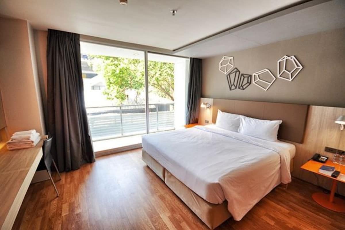 Una habitación de hotel moderna con una cama grande, un escritorio con silla, una mesita de noche y vistas al exterior a través de un amplio ventanal.