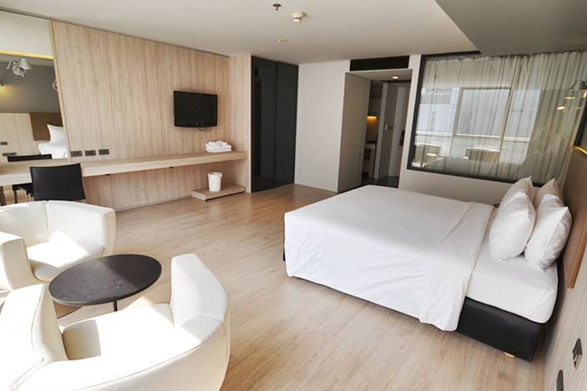 Una habitación de hotel moderna con una cama grande, una zona de estar con una mesa, un televisor y un baño visible a través de una puerta abierta.