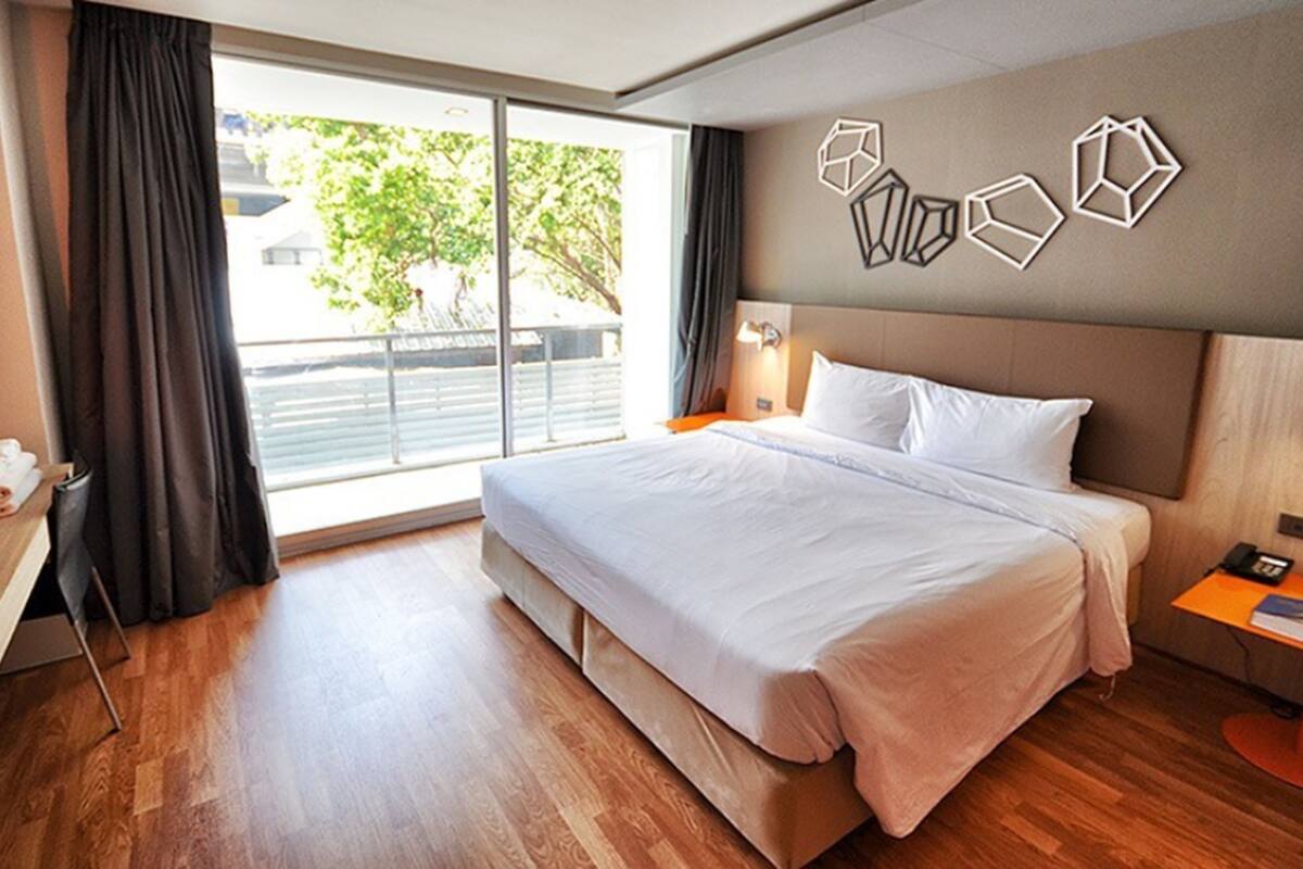 Una habitación de hotel moderna con una cama grande, suelo de madera, una ventana con cortinas, una mesita de noche con lámpara y decoración de pared geométrica.