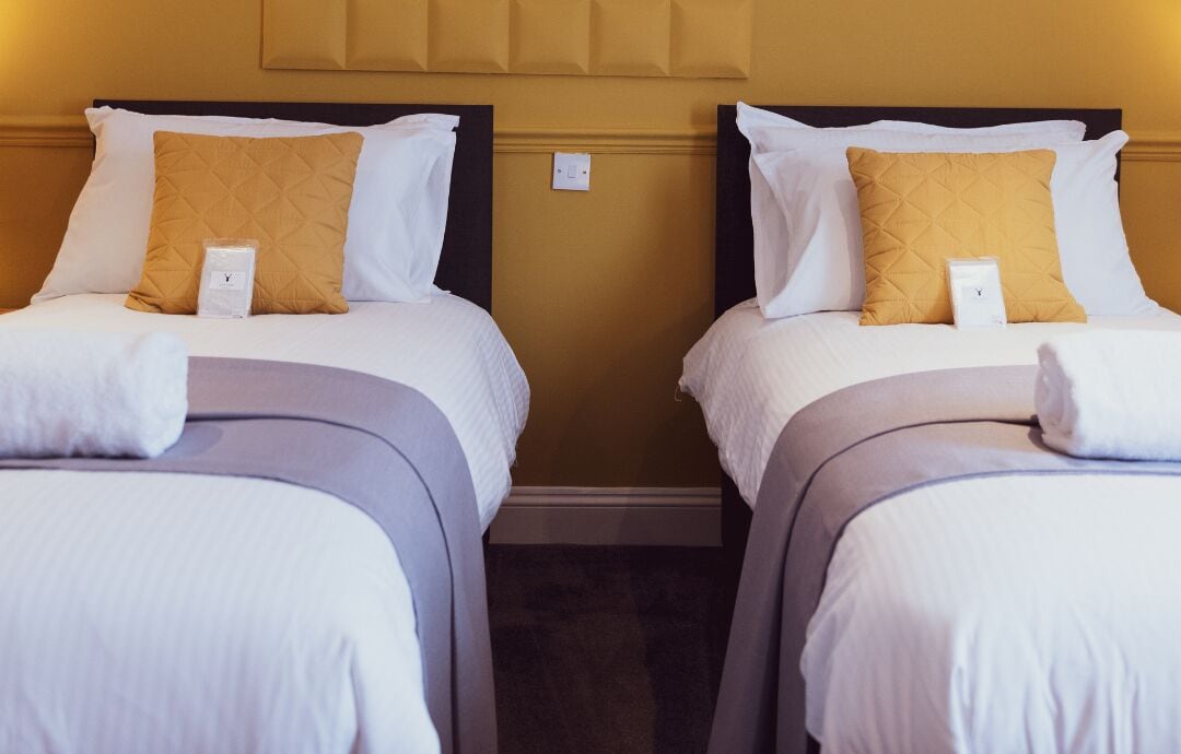 Two single beds with white and yellow pillows, a wooden headboard, and a small nightstand with a white object on each bed.
