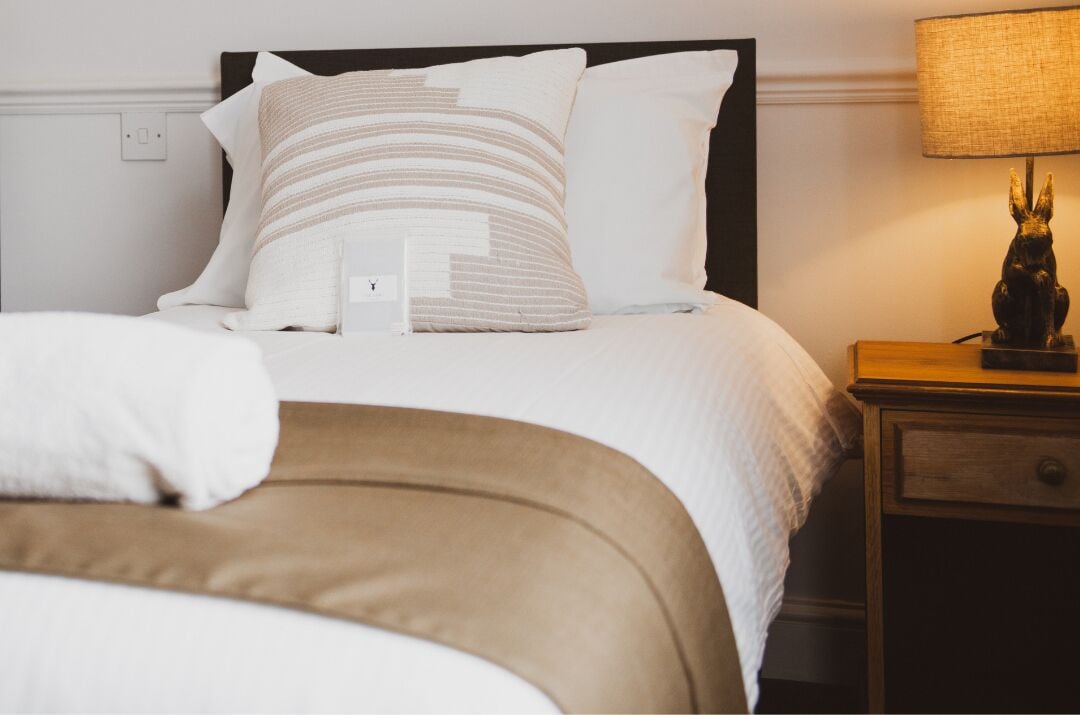 A neatly made bed with white and beige bedding, a wooden nightstand with a lamp, and a small statue.