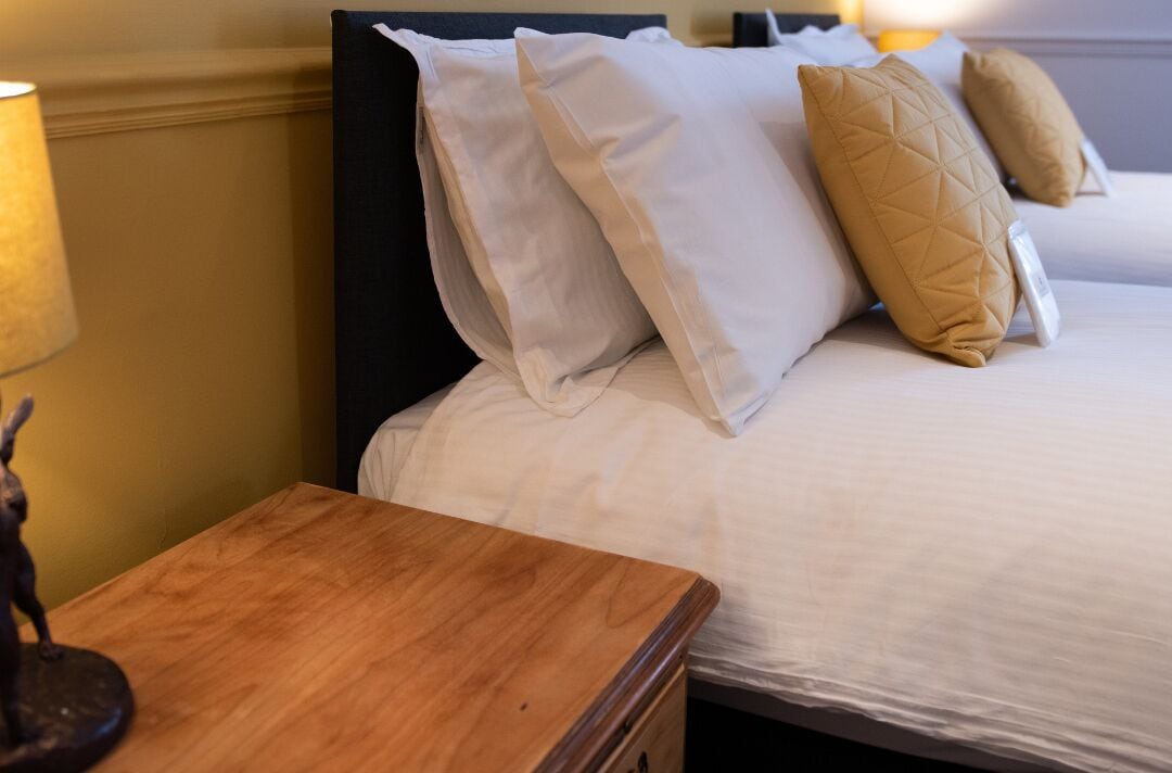 A neatly made bed with white and beige pillows, a wooden nightstand, and a lamp.