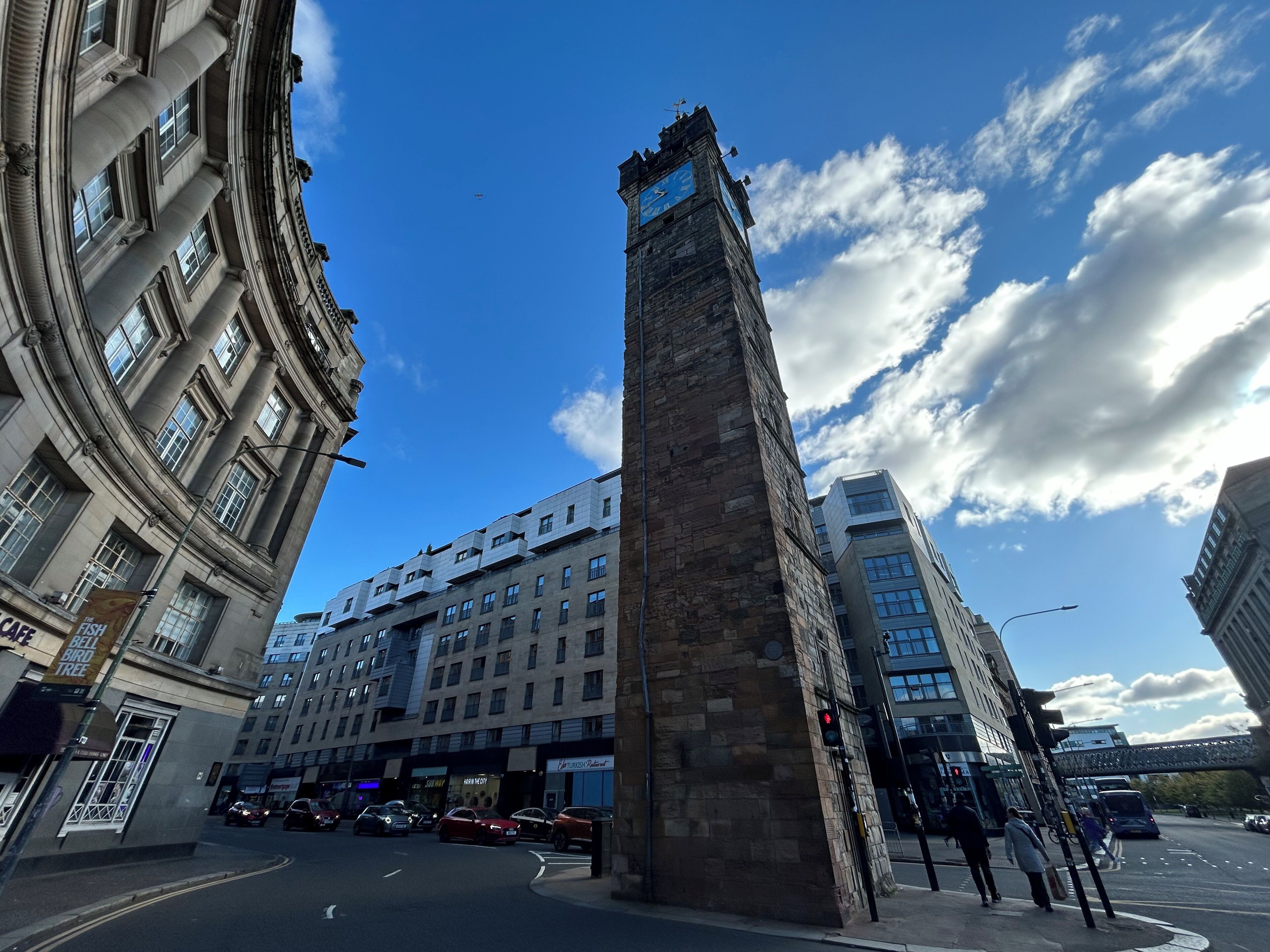 A tall stone tower with a clock at the top, surrounded by modern buildings and a busy street with cars and pedestrians.