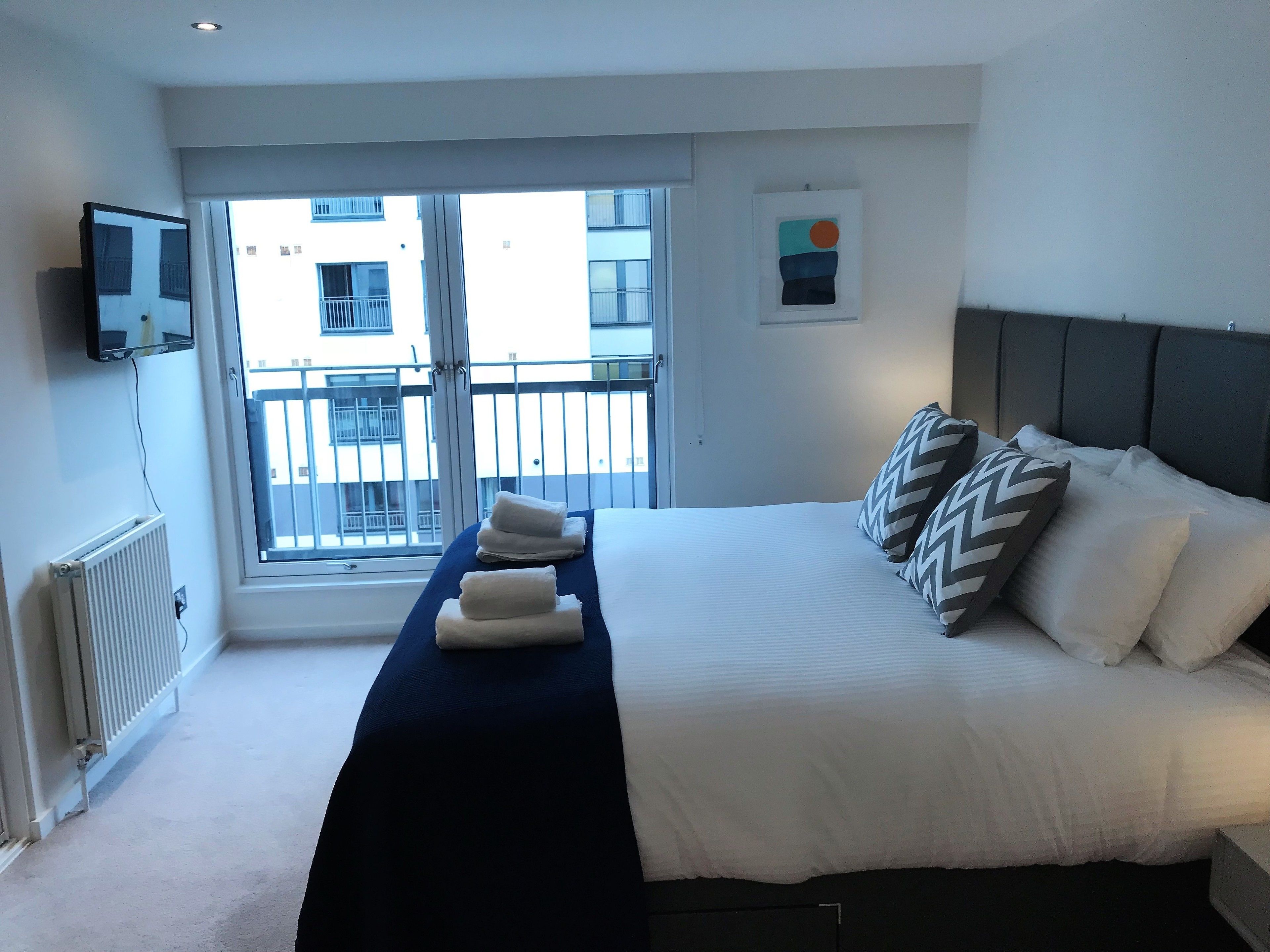 A hotel room with a bed, a TV mounted on the wall, a window with a view of buildings, and a radiator.