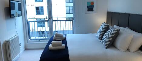 A hotel room with a bed, a TV mounted on the wall, a window with a view of buildings, and a radiator.