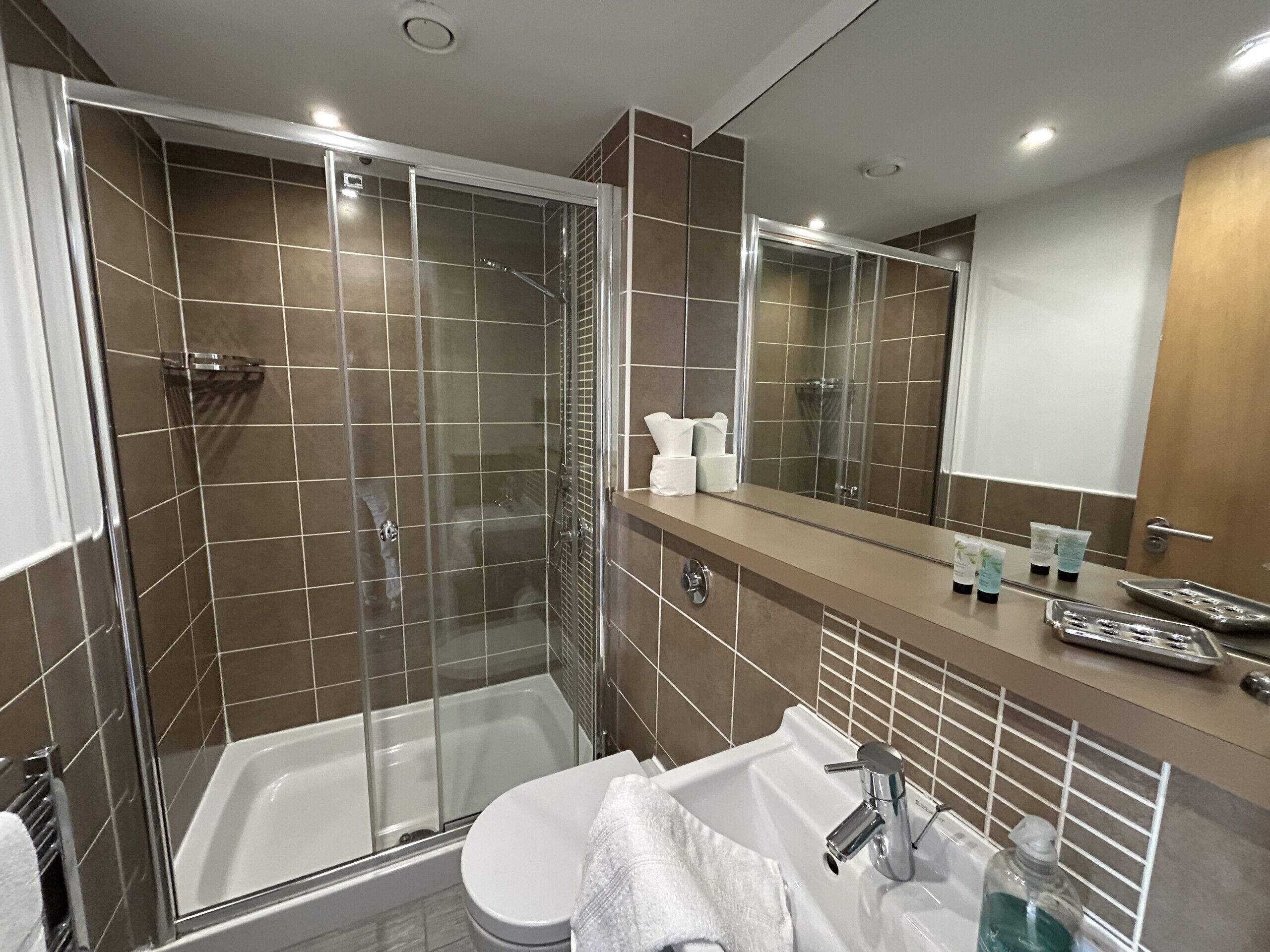 Un baño moderno con ducha de vidrio, una bañera blanca y un lavamanos con artículos de aseo.