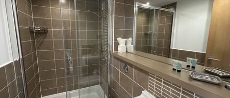Un baño moderno con ducha de vidrio, una bañera blanca y un lavamanos con artículos de aseo.