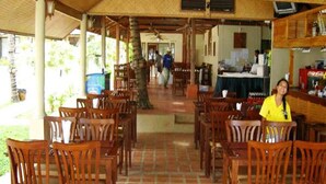 A covered outdoor dining area with wooden tables and chairs, a Coca-Cola refrigerator, and a view of the outdoor space. - Samui Beach Apartments (Koh Samui)