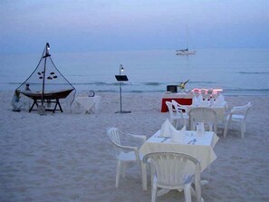 Un montaje para comer frente al mar con mesas y sillas, un bote pequeño y un velero a lo lejos.
