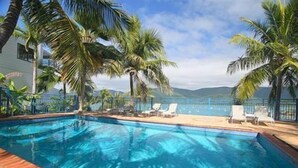 A swimming pool with palm trees and lounge chairs, surrounded by a fence and overlooking a body of water. - Coral Point Lodge (Shute Harbour)