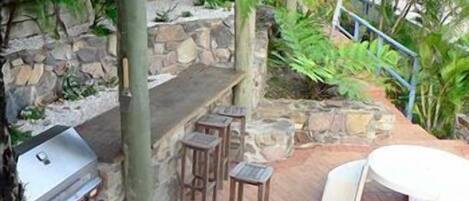 An outdoor patio with a stone wall, a grill, wooden stools, and a white table.