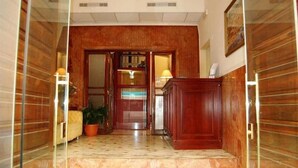 Un vestíbulo de hotel con suelo de mármol, paneles de madera, un mostrador de recepción y una planta en maceta.