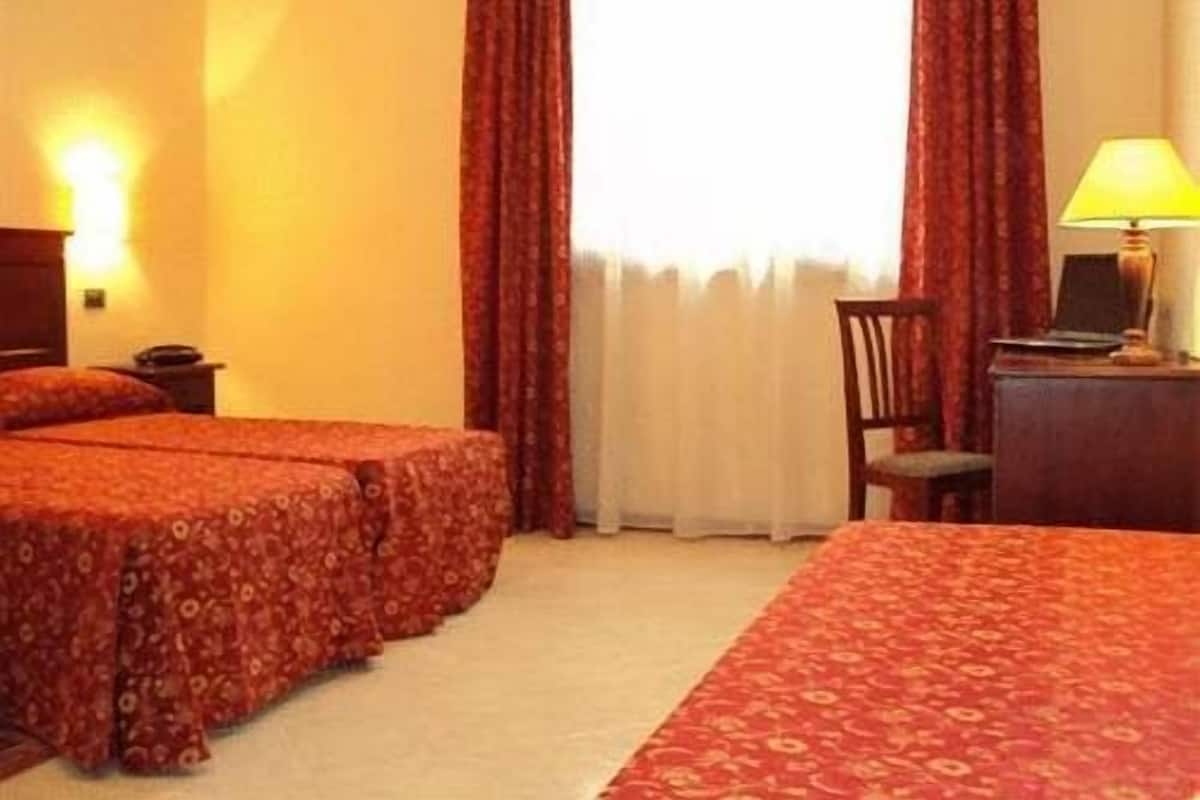 A hotel room with two beds, a desk with a lamp, and a view of the outdoors through a window with curtains.