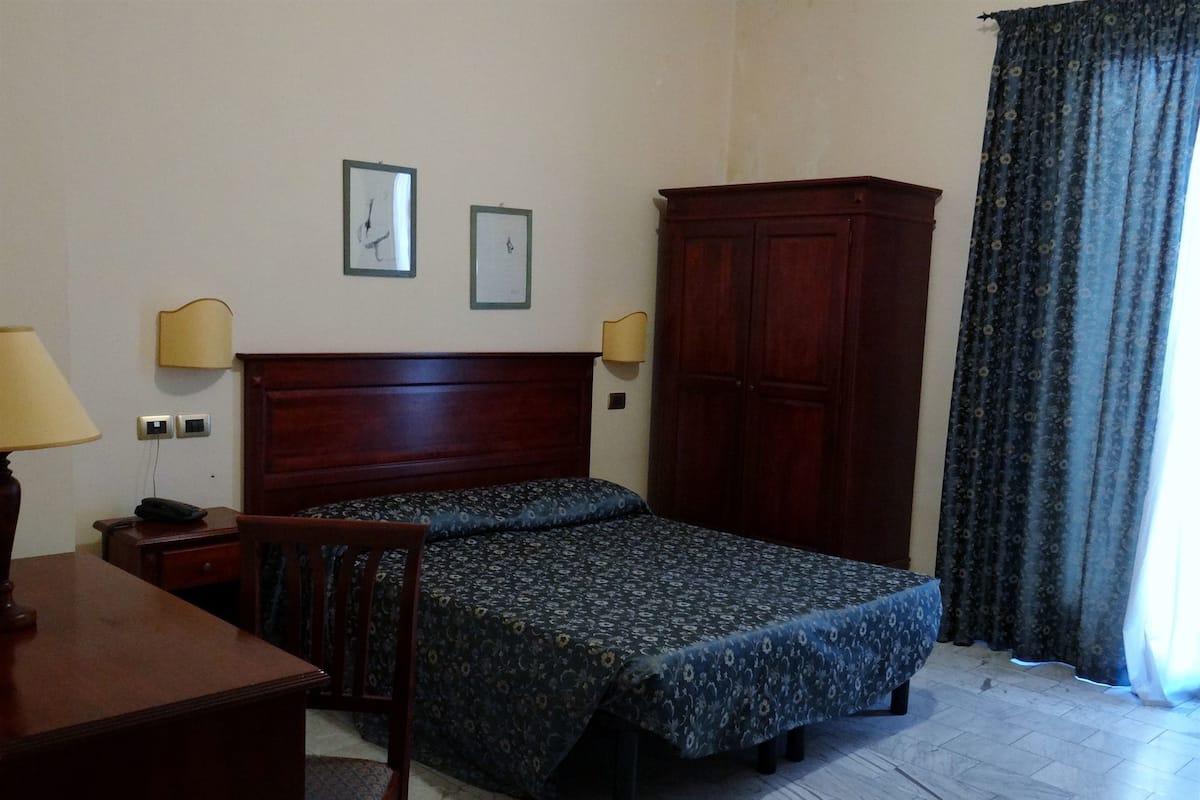 A bedroom with a bed, a wooden wardrobe, a desk with a lamp, and two wall clocks.