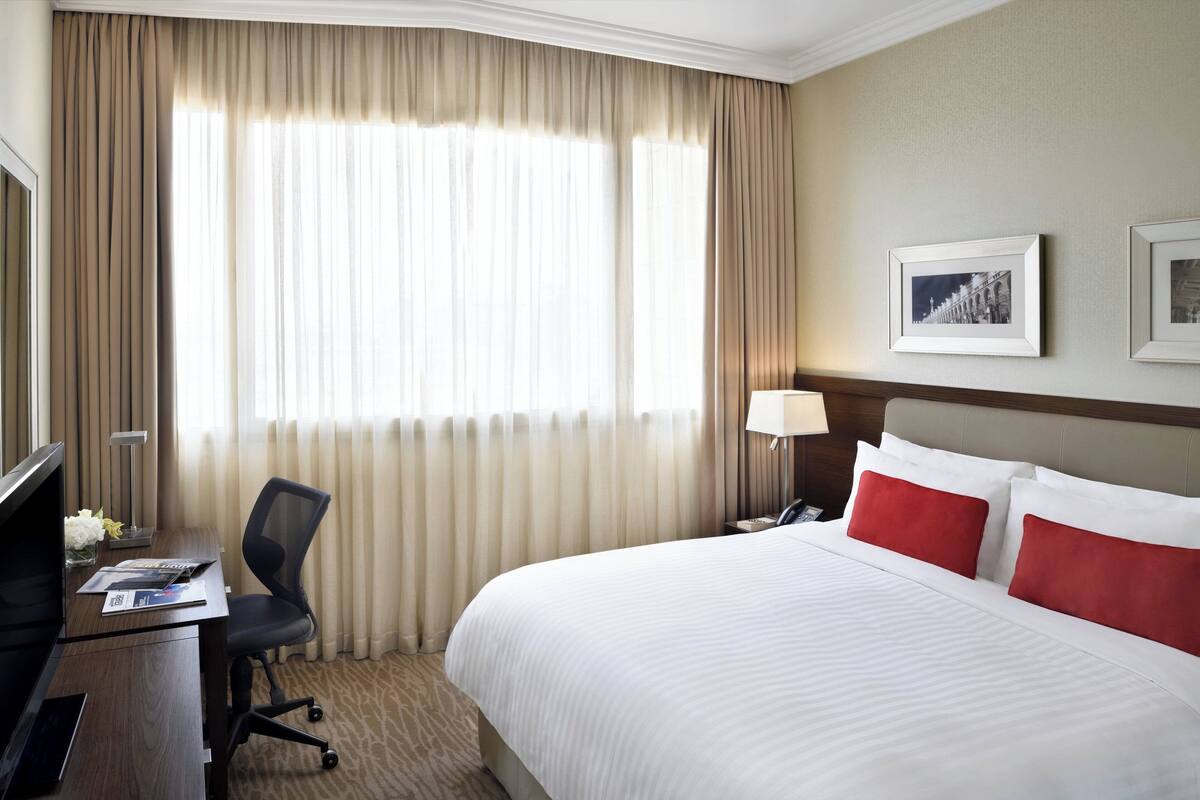 A hotel room with a large bed, a desk with a chair, a lamp, and two framed pictures on the wall.