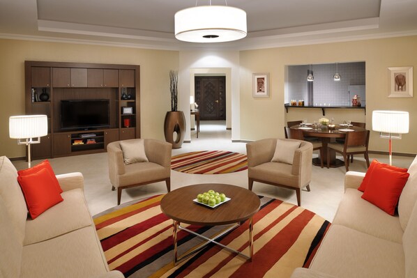 A modern living room with a flat-screen TV, two beige armchairs, a coffee table with a fruit bowl, and a striped rug.