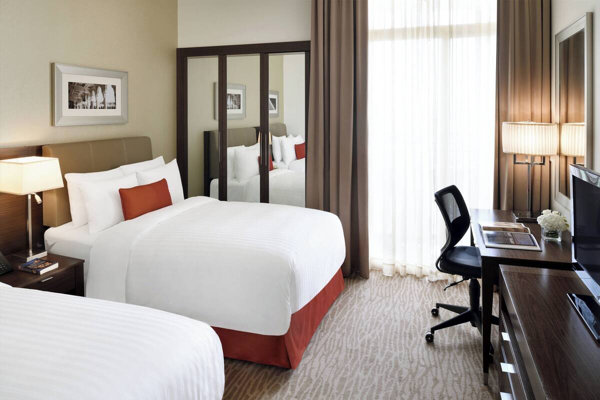 A hotel room with two beds, a desk with a chair, a TV, and a lamp.