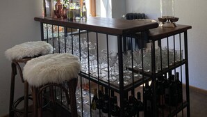 Zona de bar con mostrador de madera, dos taburetes con cojines blancos y mullidos, y estanterías con botellas.