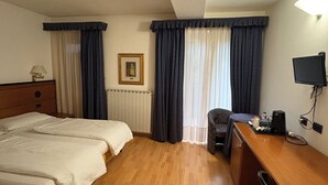 Habitación de hotel con dos camas, un escritorio, una silla, un radiador, un cuadro en la pared y un televisor en la pared.