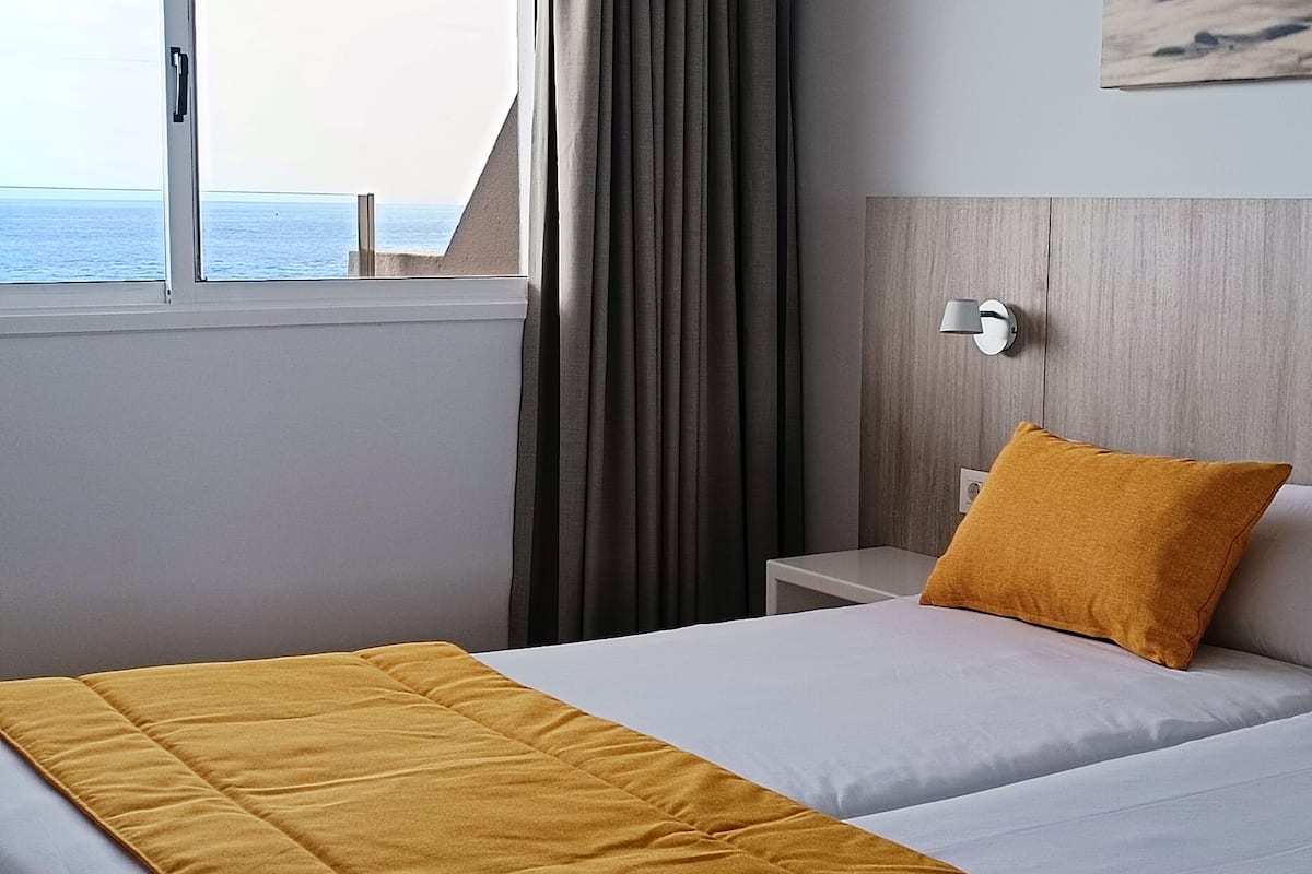 A hotel room with a bed, a nightstand, a lamp, and a view of the sea through a window.