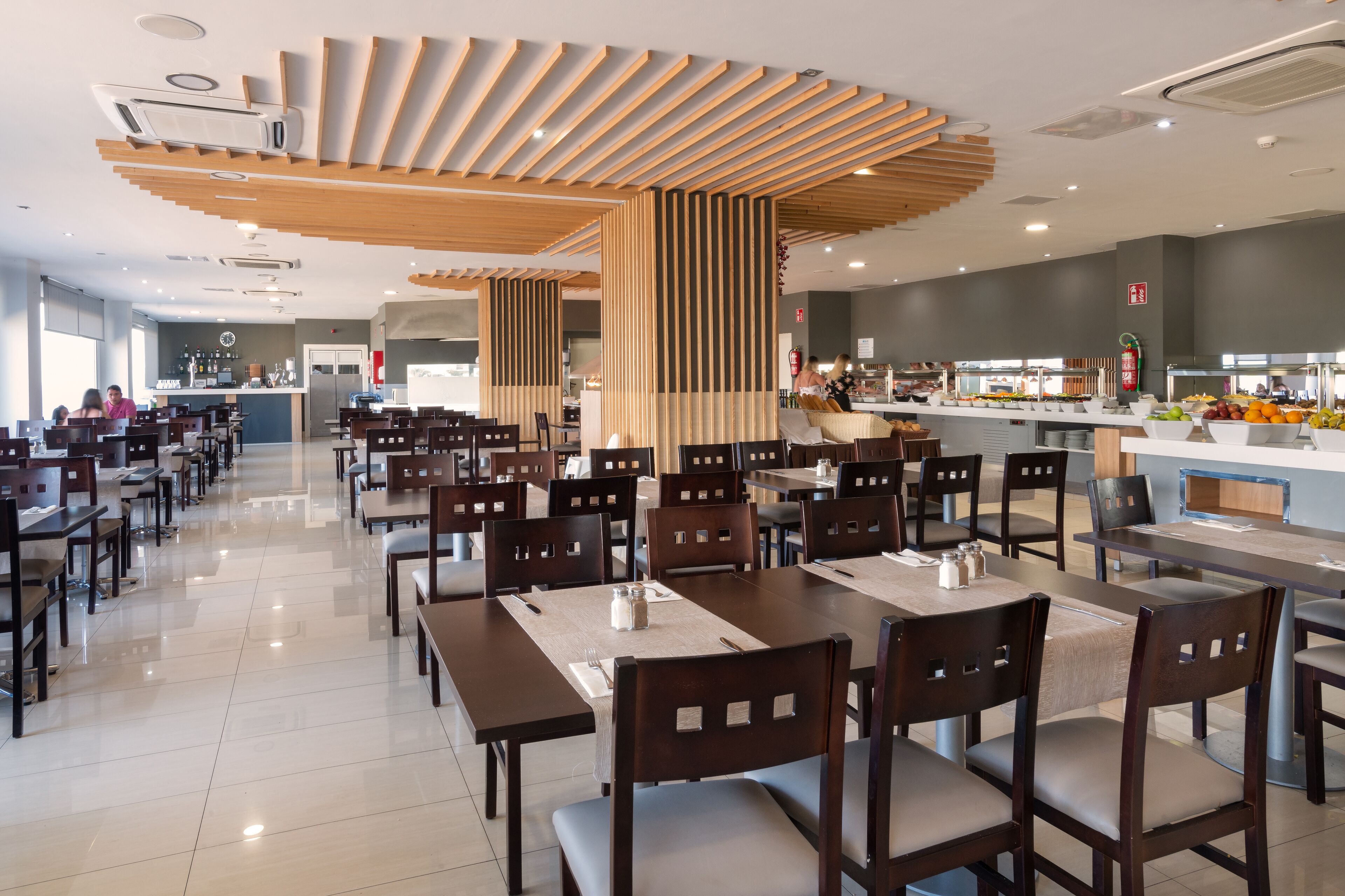 A spacious cafeteria with wooden tables and chairs, a central serving area, and a clean, modern design.