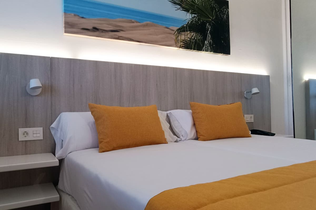 A hotel room with a bed, white bedding, two orange pillows, a nightstand, a wall-mounted lamp, and a painting of a beach scene.