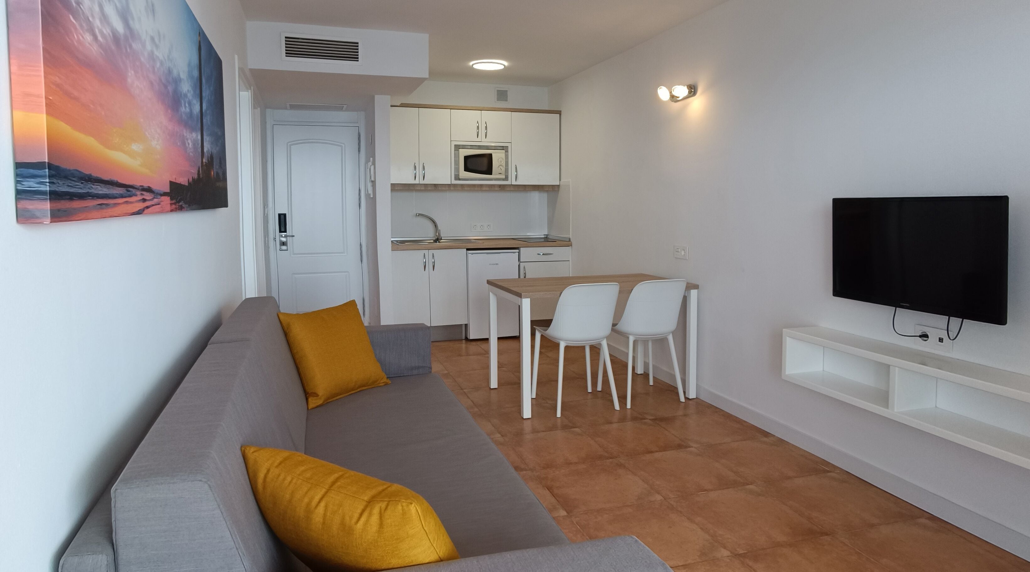 A modern living room with a gray sofa, a wooden dining table, white chairs, a flat-screen TV, and a kitchen in the background.
