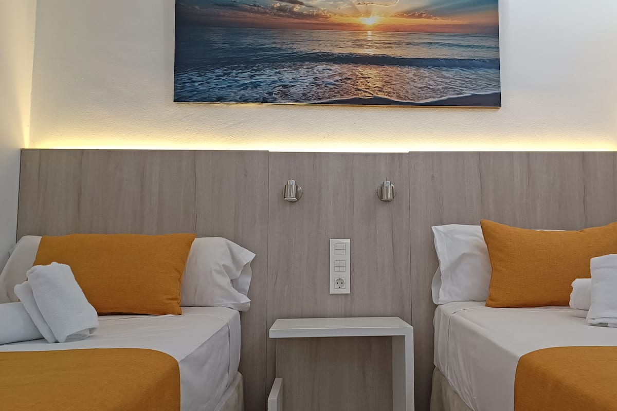 A hotel room with two beds, each with white linens and orange pillows, a small bedside table, and a wall-mounted light.