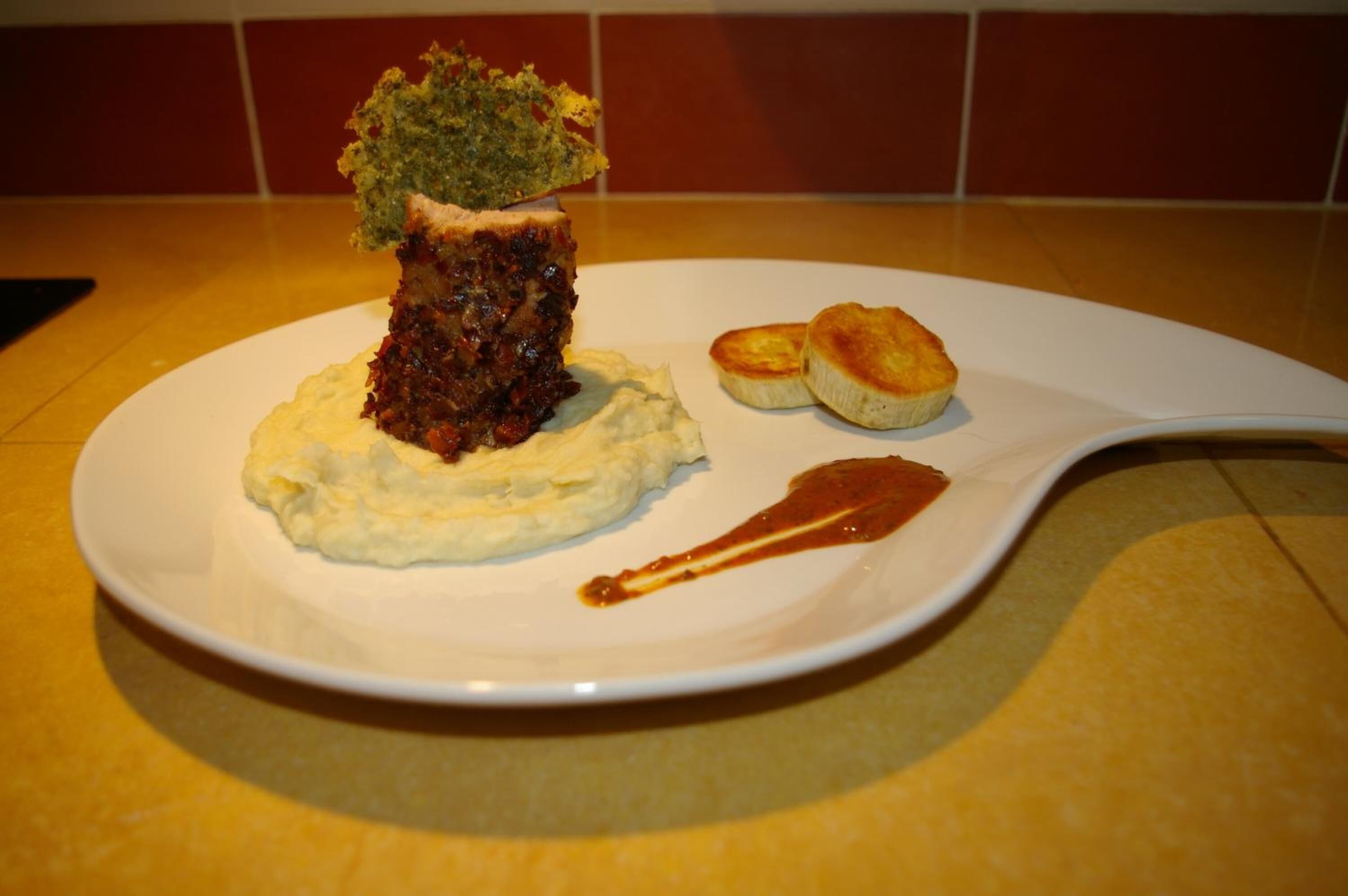 Un plato blanco con puré de papas, un trozo de carne y dos pequeños pasteles sobre una mesa amarilla.
