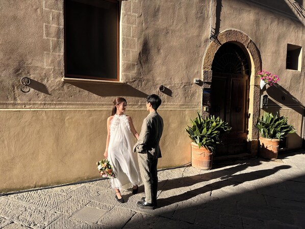A couple walking past a building with a stone facade, a wooden door, and potted plants.