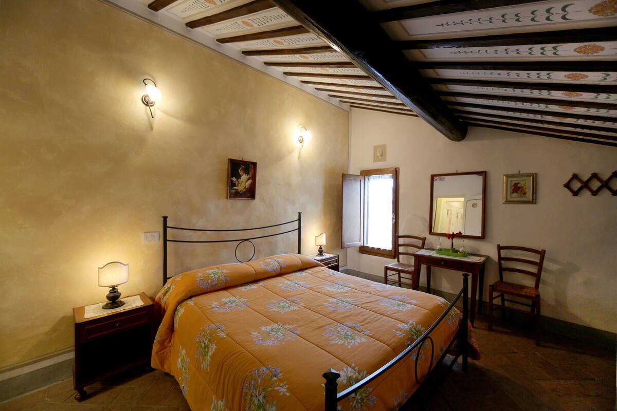 A bedroom with a bed, nightstand, lamp, chair, table, and a mirror.