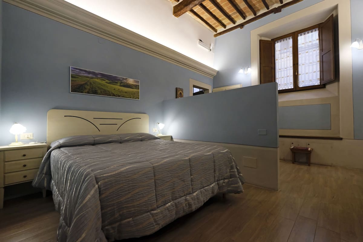 A bedroom with a bed, bedside lamps, a nightstand, a window with curtains, and a painting on the wall.