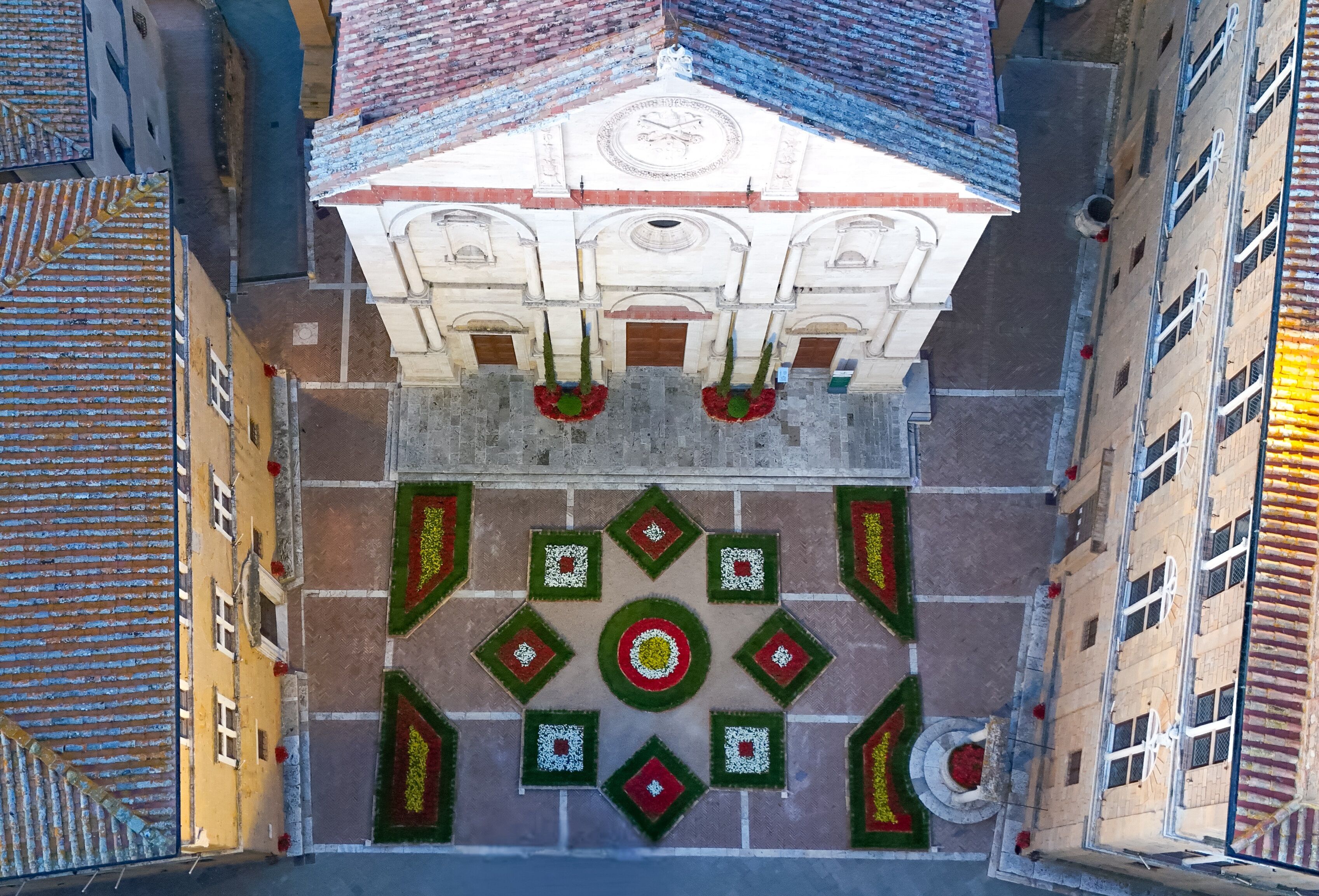 Vista aérea de una plaza con una fuente central, rodeada de edificios con tejados de azulejos y una torre del reloj.