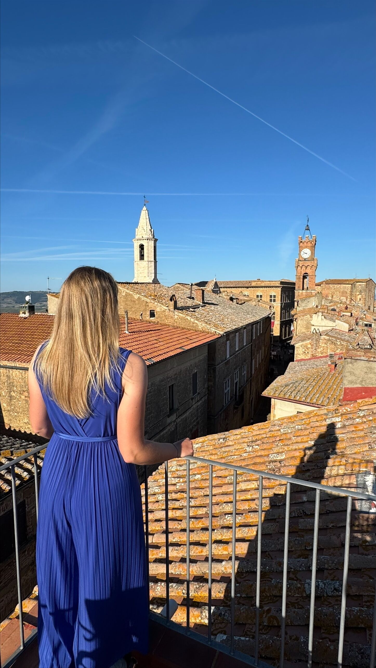 A woman in a blue dress standing on a balcony overlooking a historic town with tiled roofs and two prominent towers.