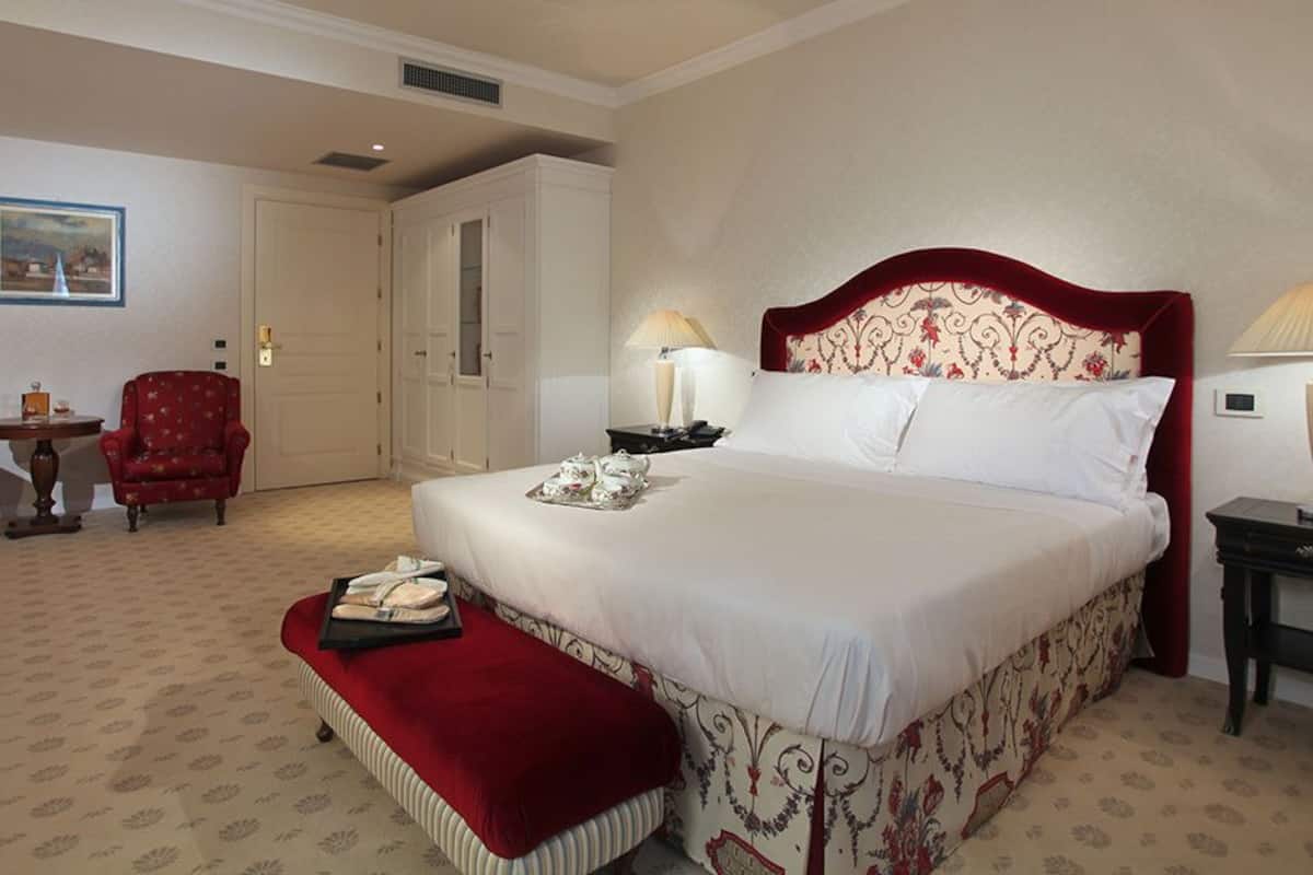 A hotel room with a large bed, two armchairs, a small table, and a red bench.