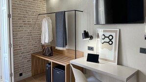 A modern bathroom with a brick wall, a wooden bench, a towel rack, a black bag, a framed picture, a mounted TV, and a wall-mounted light fixture. - Cosy Rooms Embajador (Valencia)